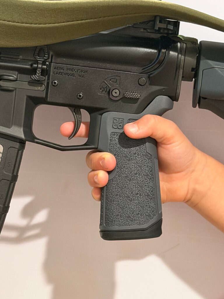 A young shooter's hand on a standard full-size AR grip: the fingers claw at the front of the grip and only the fingertip reaches the trigger